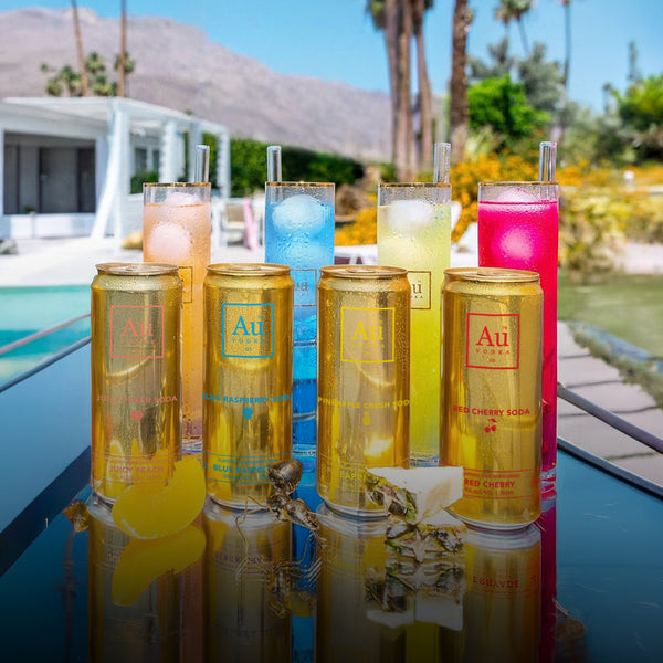 Four Au Vodka soda cans and four cocktails on a table by a pool with mountains in the background